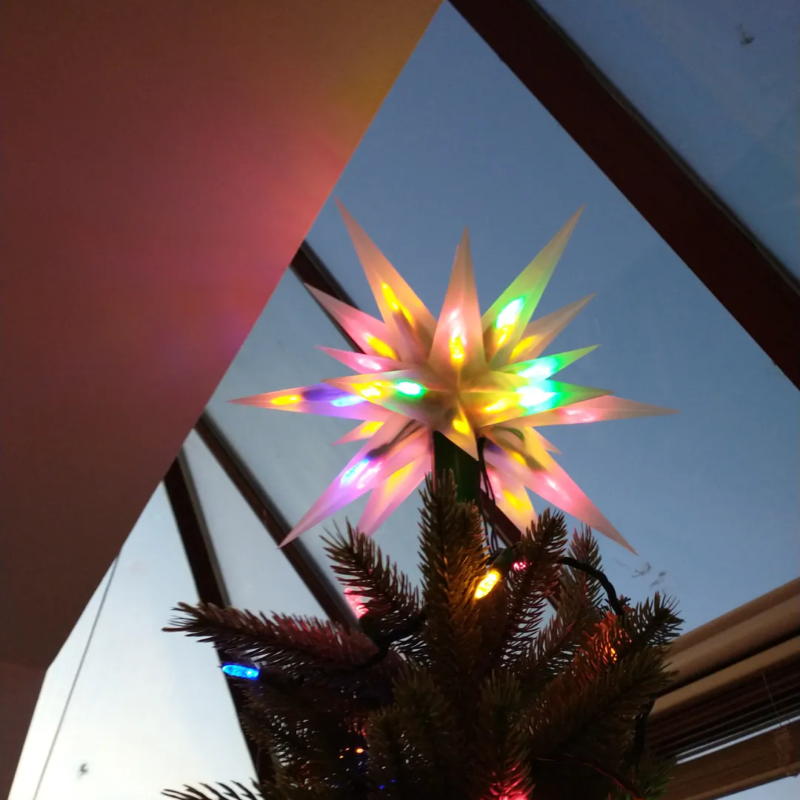 There is a colorful lighted Moravian star tree topper on a Christmas tree